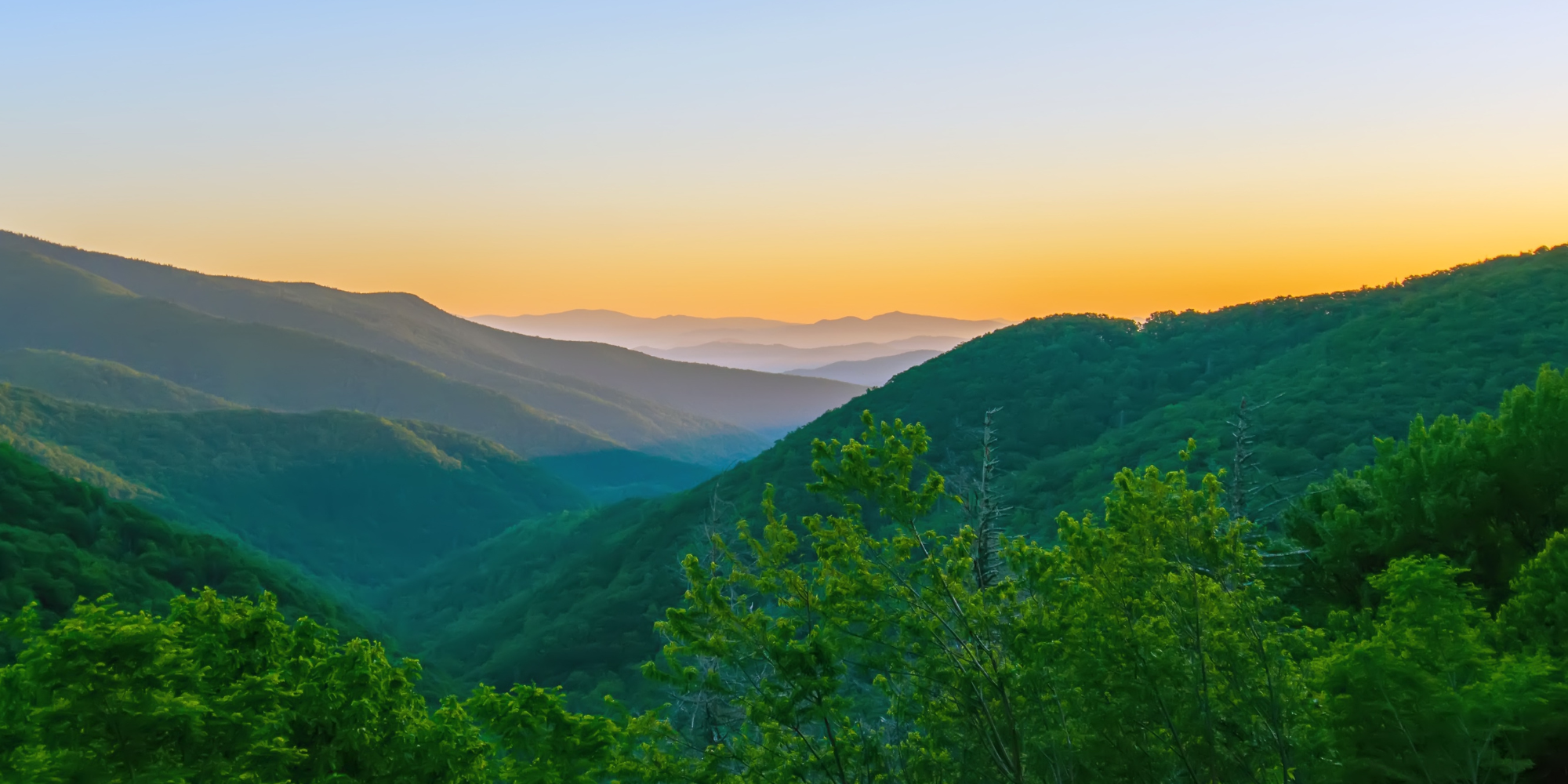 Sunset over lush green mountains with layers of hills fading into the distance. The sky is a gradient of orange and blue, creating a peaceful atmosphere.